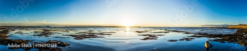 Panorama of Rimouski sunset in Gaspesie region of Quebec, Canada with sun path, seaweed, rocks and shallow water, reflection