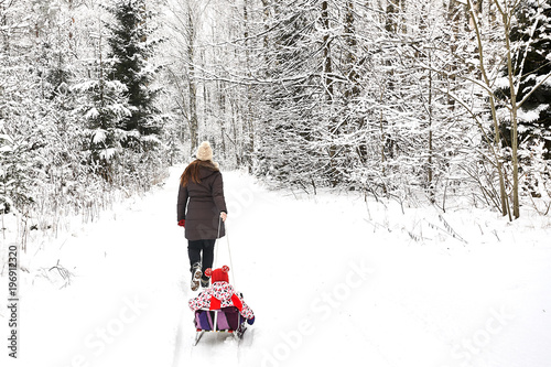 Young beautiful girl whith wihite hat  posing against a winter park background and playing with the girl child with the snow in forest. Towing sledge