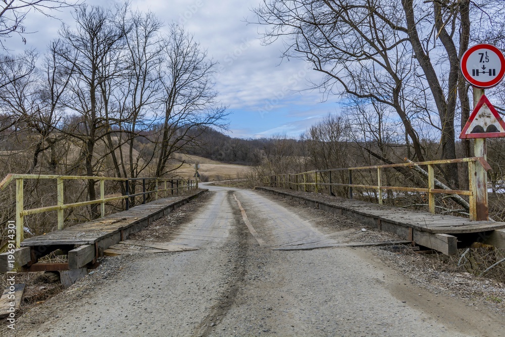 Wooden single lane bridge with weight restriction road sign and bumpy road sign. Old wooden bridge over the river in winter season. Wooden single lane bridge with weight restriction road sign and bumpy road sign. Old wooden bridge over the river in winter season.
