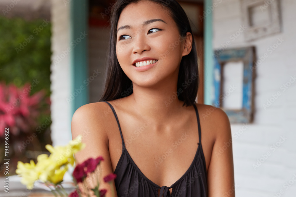 Positive smiling satisfied Chinese female with healthy skin dressed casually, looks happily away, sits against home domestic interior enjoys good weather out of window. Lovely Asian woman poses indoor Positive smiling satisfied Chinese female with healthy skin dressed casually, looks happily away, sits against home domestic interior enjoys good weather out of window. Lovely Asian woman poses indoor