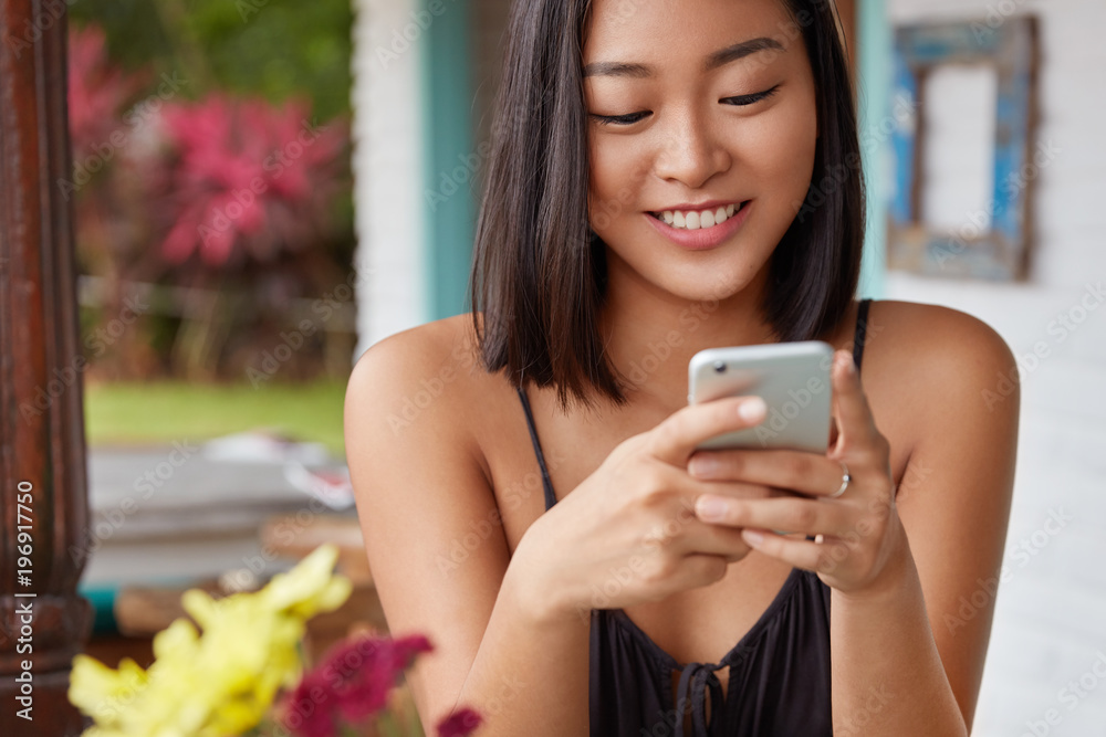 Cropped shot of happy Japanese female holds modern smart phone, enjoys recreation in cafe, messages in social networks during evening rest. Cheerful young Asian woman sends photo via cellular Cropped shot of happy Japanese female holds modern smart phone, enjoys recreation in cafe, messages in social networks during evening rest. Cheerful young Asian woman sends photo via cellular