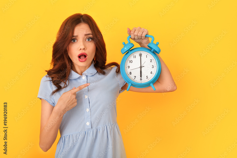 Young woman studio isolated on yellow holding alarm clock pointing at time - obrazy, fototapety, plakaty 
