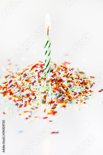 Burning white and green striped spiral birthday candle for dopping cakes, cupcakes or muffins as decoration on pile of colorful sugar spinkles on white background photo