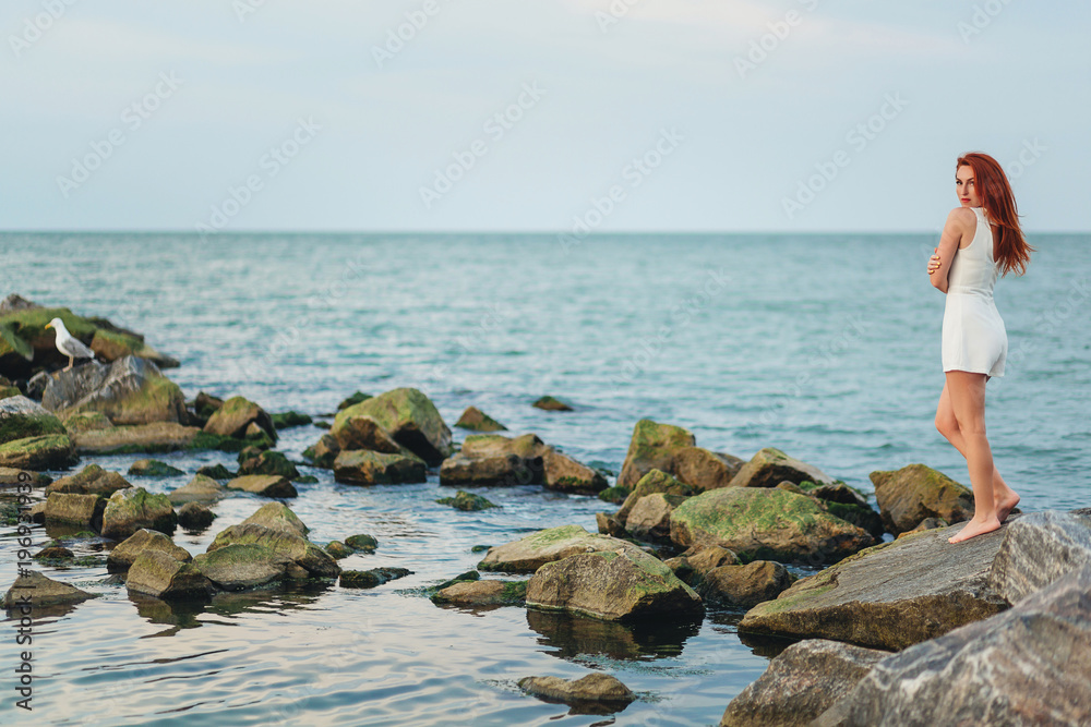 Young pretty redhead woman stand alone on the rocks near the sea