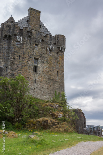 Dornie, Scotland - June 10, 2012: Gray-brown-stone main house and tower of Eilean Donan Castle. Green vegetation up front. Gray cloudscape and one artillery piece.