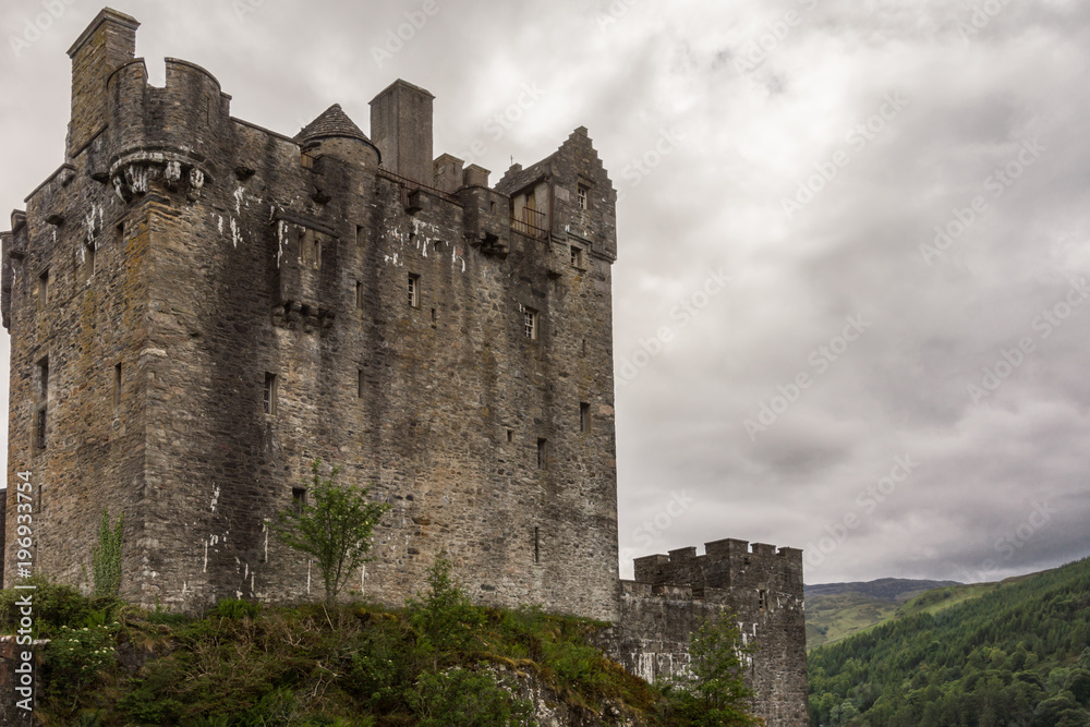 Dornie, Scotland - June 10, 2012: Gray-brown-stone main house and tower of Eilean Donan Castle. Green vegetation up front. Gray cloudscape . Mountains as backdrop.