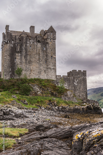 Dornie, Scotland - June 10, 2012: Gray-brown-stone main house and tower of Eilean Donan Castle. Green vegetation on top of rocky loch shore up front. Gray cloudscape . Mountains as backdrop.
