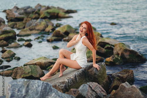 young redhead woman sit on rocks and laughs near the ocean