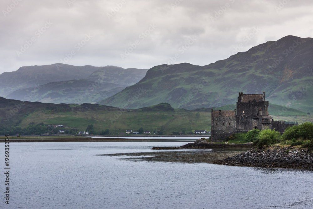 Dornie, Scotland - June 10, 2012: Brown-stone Eilean Donan Castle with the confluence of  Loch Duich, Loch Long and Loch Alsh. Green forested hills as backdrop. Rain-heavy cloudscape.