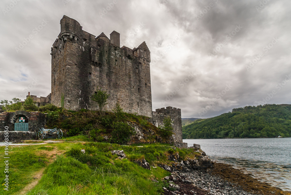 Dornie, Scotland - June 10, 2012: Brown and grey stone main house and tower of Eilean Donan Castle. Green vegetation up front and gray cloudscape. War memorial with artillery piece.
