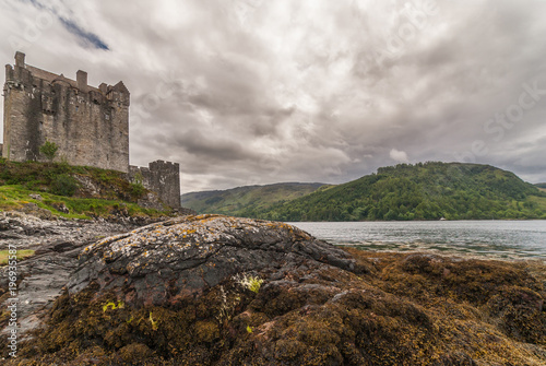 Dornie  Scotland - June 10  2012  Brown and grey stone main house and tower of Eilean Donan Castle off center. Rock and brown seaweed up front . Gray cloudscape. Loch and green hills on horizon.