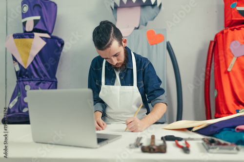 Male designer in the craft workshop working. Young designer drawing on the desk with a laptop and tools