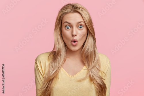 Картината върху платно Isolated shot of pretty young female stares at camera with shocked expression, recieves unexpected news, keeps lips rounded, poses in studio over pink blank wall