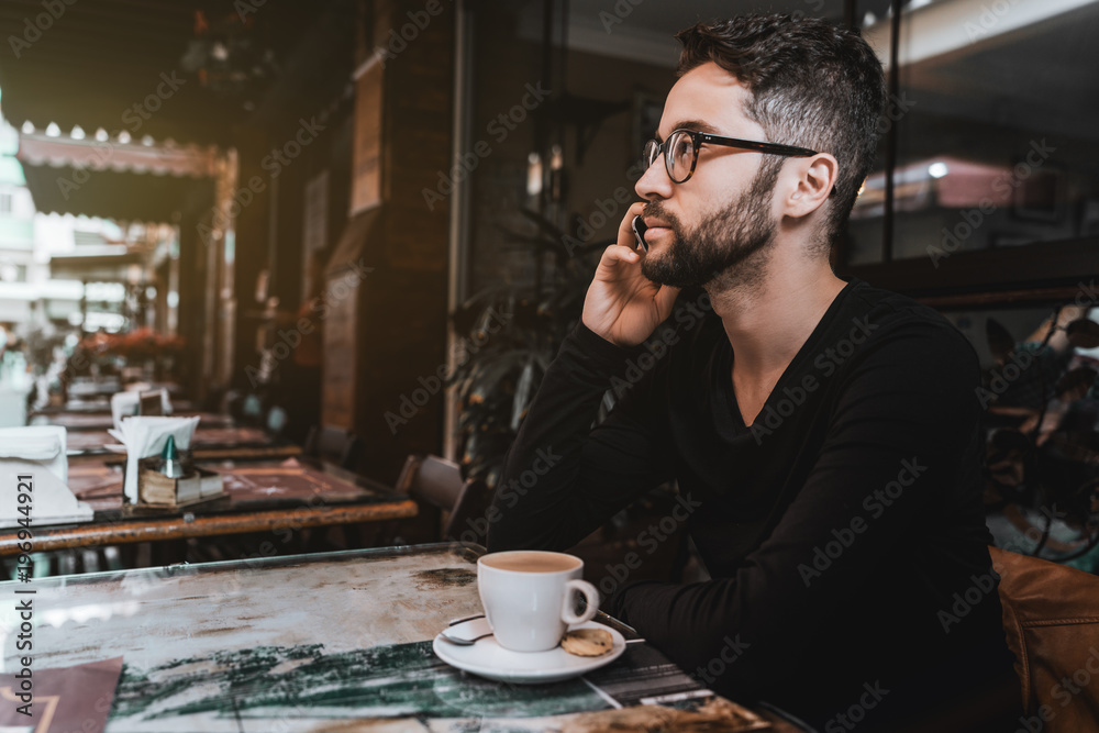 Young good-looking hipster guy with the beard and in glasses is talking on the phone with his colleague while sitting in a street bar outdoor and having a breakfast with the cup of a tasty coffee