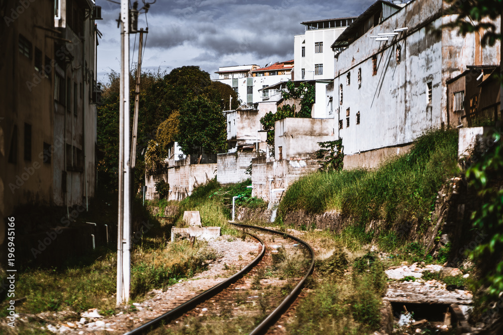 View of a railway track overgrown with grass stretching between residential houses and favelas, and turning to the left in the distance, bright summer day, overcast sky, Juiz de Fora, Brazil
