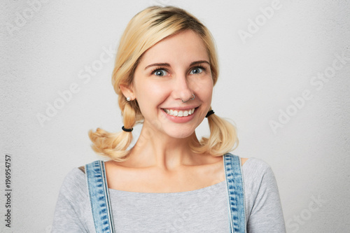 Indoor shot of young blonde girl with perced nose, wears overalls, simply hairdo smiling and show her teeth to the world. Isolated over white concrete wall photo