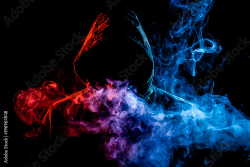 A young man in a black hood smokes a vape and exhales a colored blue, pink, yellow smoke on a black isolated background