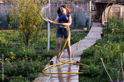 Sensual caucasian woman pours water from a hose flowers and vegetables, trees in her garden. A hot summer day, a river in the background, a greenhouse, an excellent harvest of fruits and vegetation. photo