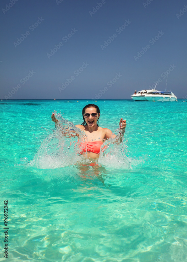 Sunglasses wearing young woman in crystal clear sea water splashing water in front of her with smiling happy face  expression. Similan Islands, Thailand