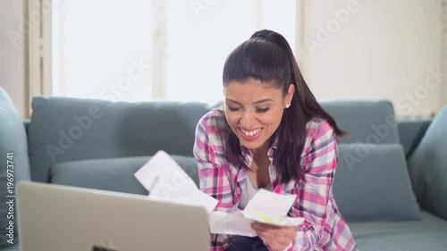 lateral pan shot of young attractive happy hispanic woman accounting payments and calculating monthly expenses bills bank papers and paperwork smiling at apartment couch in domestic finance success