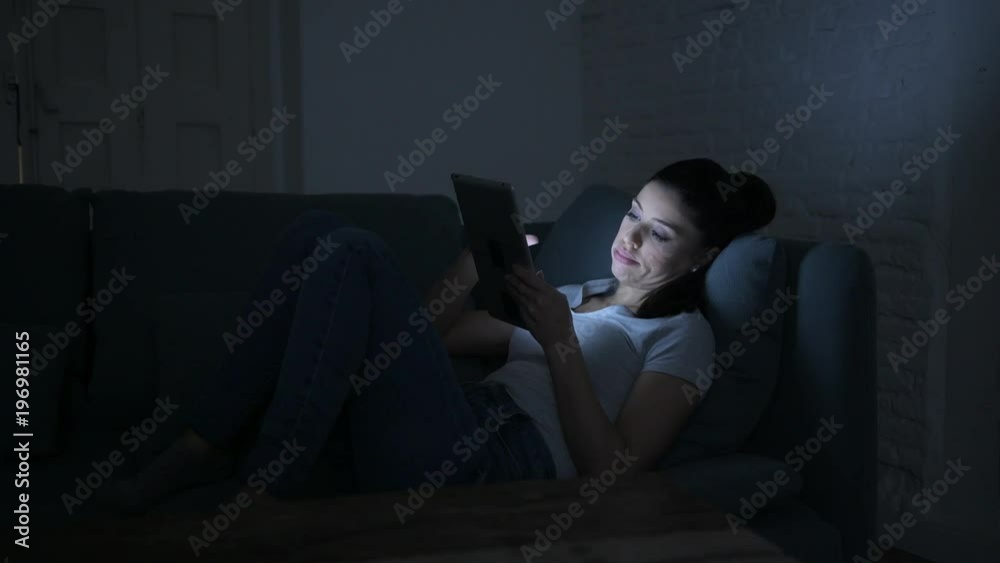 lateral pan shot of young beautiful happy and relaxed latin woman lying on home couch late night using digital device laptop tablet pad watching internet movie lateral pan shot of young beautiful happy and relaxed latin woman lying on home couch late night using digital device laptop tablet pad watching internet movie