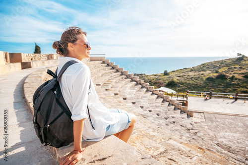 Billede på lærred A young beautiful woman traveler sits on the steps of an amphitheater in an architectural antique museum overlooking the sea