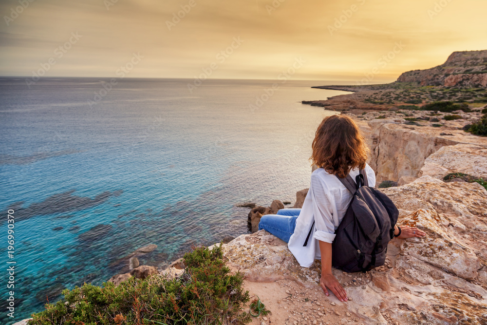 A stylish young woman traveler watches a beautiful sunset on the rocks on the beach, Cyprus, Cape Greco, a popular destination for summer travel in Europe - obrazy, fototapety, plakaty 