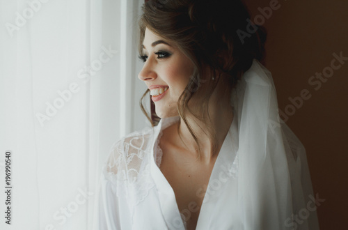 Young gorgeous bride is smiling near the window waiting for her groom. Tender light and tulle veil. Beautiful photo of bridal morning. Black and white.