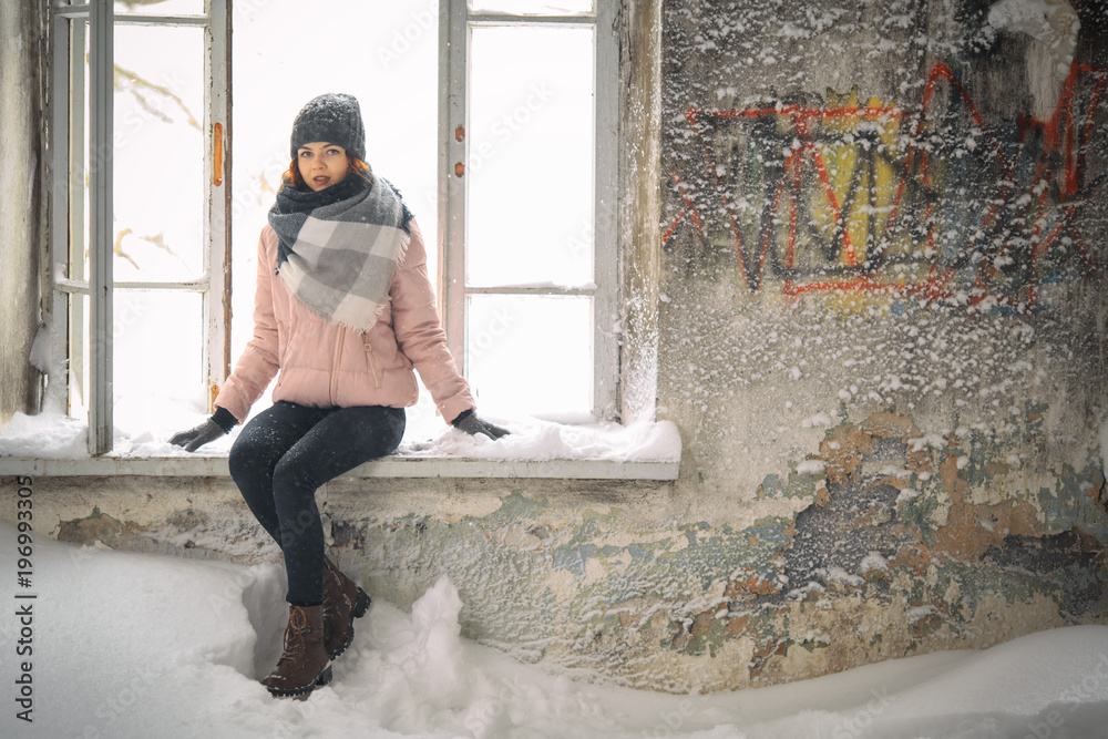A young cute white girl in a hat and warm shawl is sitting on the windowsill of a ruined house in winter.