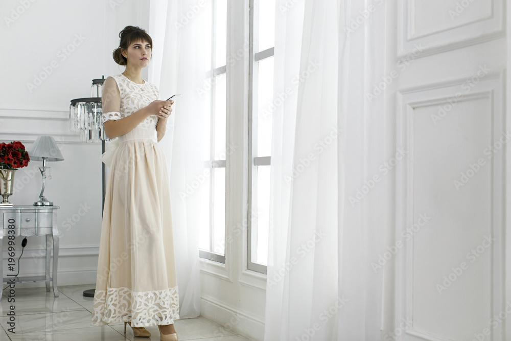 Young beautiful girl in a light long dress near a large window Young beautiful girl in a light long dress near a large window