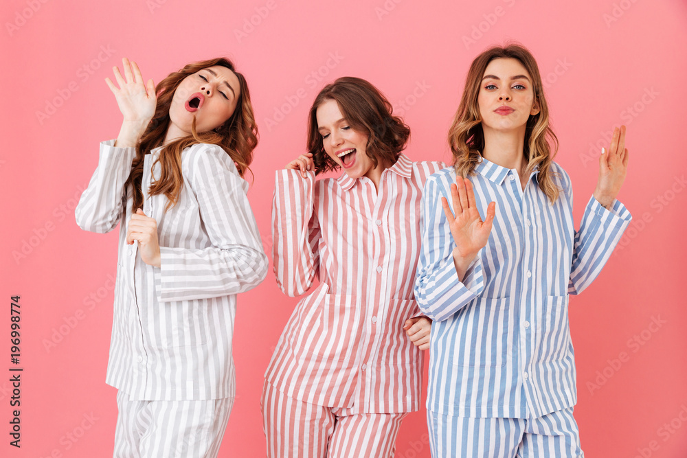 Portrait of three beautiful young girls 20s wearing colorful striped pyjamas having fun during sleepover, isolated over pink background - obrazy, fototapety, plakaty 