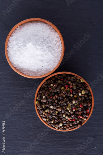 Conceptual composition of salt and pepper on spoons and bowls over dark background, top view, close-up