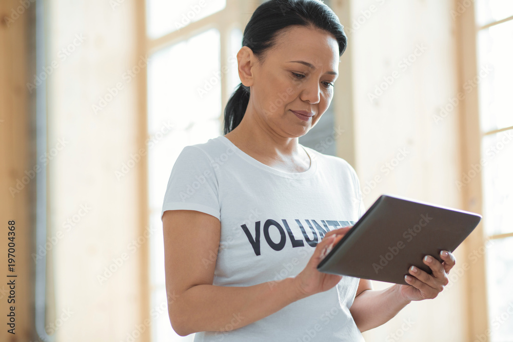 Modern device. Low angle of pleasant earnest female volunteer standing on blurred background and carrying tablet while staring down Modern device. Low angle of pleasant earnest female volunteer standing on blurred background and carrying tablet while staring down