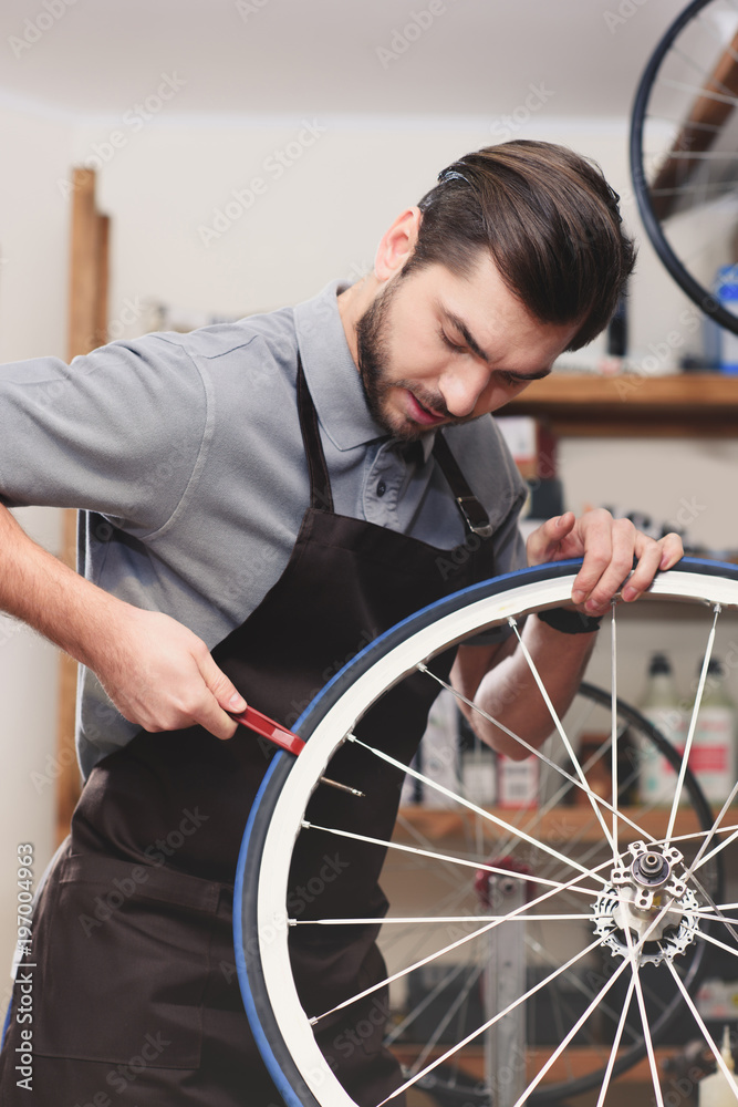 handsome young worker in apron fixing bicycle wheel in workshop