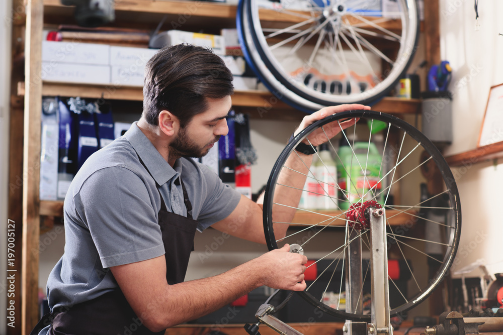 young man in apron repairing bicycle wheel in workshop
