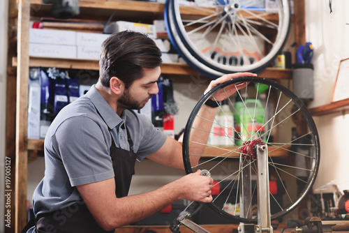 Fototapeta Naklejka Na Ścianę i Meble -  young man in apron repairing bicycle wheel in workshop