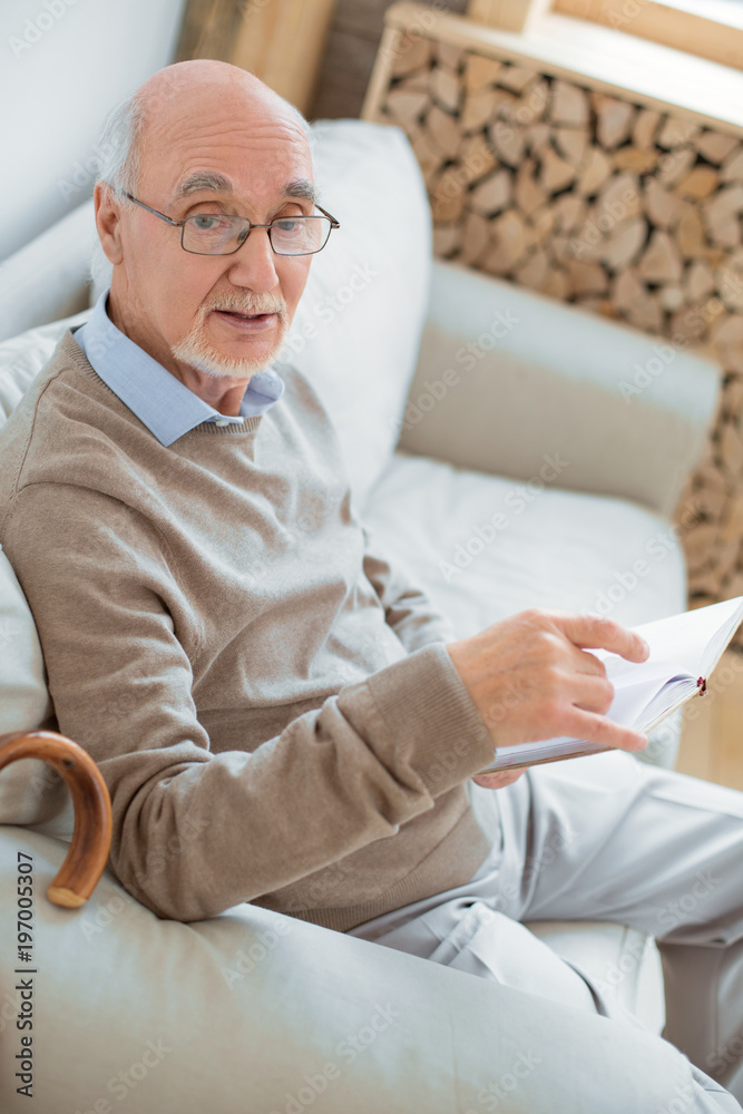 Ordinary day. Top view of smart intelligent senior man sitting on sofa while holding book and wearing glasses Ordinary day. Top view of smart intelligent senior man sitting on sofa while holding book and wearing glasses