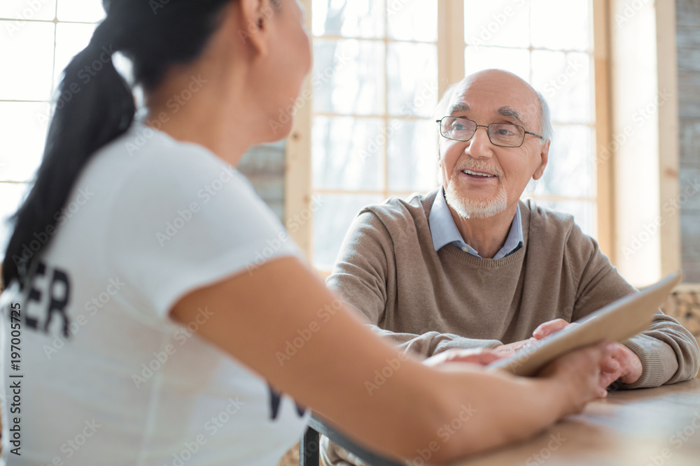 Fototapeta premium Twisted plot. Cheerful merry senior man laughing while listening to volunteer and sitting on blurred background