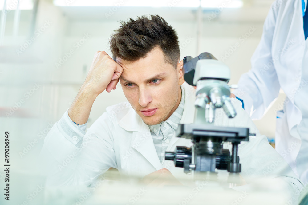 Discontent young researcher wearing white coat sitting at laboratory bench and leaning head on hand while thinking over unsuccessful experiment, his female colleague trying to encourage him Discontent young researcher wearing white coat sitting at laboratory bench and leaning head on hand while thinking over unsuccessful experiment, his female colleague trying to encourage him