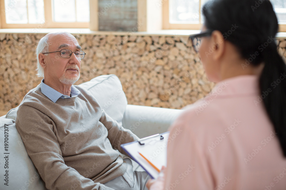 Obraz premium First impression. Charming handsome senior man sitting on sofa while looking at caregiver and communicating with her