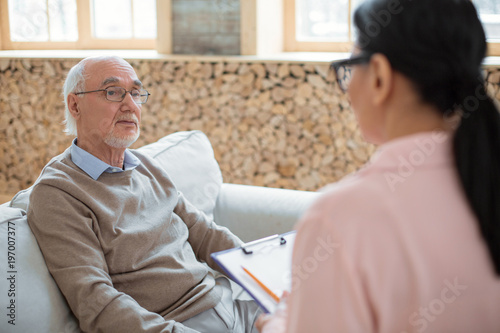 First impression. Charming handsome senior man sitting on sofa while looking at caregiver and communicating with her