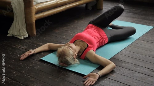 Mature woman is having yoga training in her beedroom. Blond lady with slim body is lying on wooden floor on blue mat and doing leg twists to stretch her musculs and take care of her spiritual and