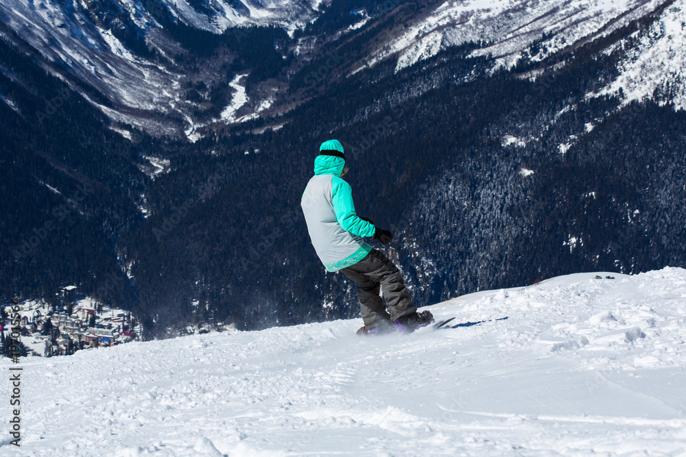 A snowboarder in a mask and gear roll on the board down the hill with his back to the camera. Below you can see the mountain forest and the snow-covered valley