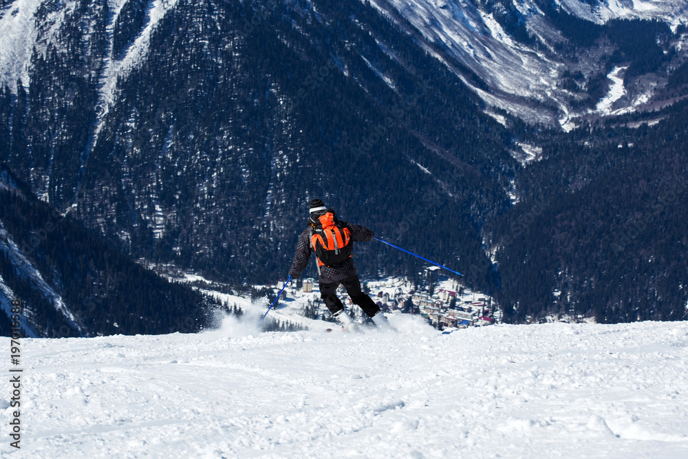 snowboarder in a mask and gear roll on the board down the hill with his back to the camera. Below the mountain forest and the snow-covered valley