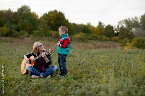 A boy and his mother are playing and singing guitar in the autumn forest