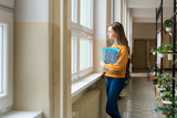 Young attractive female high school student standing by the window in the hallway at her school alone. Education concept.