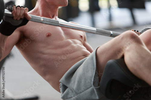 A shirtless young muscular man working out in gym doing exercises with equipment in fitness center.