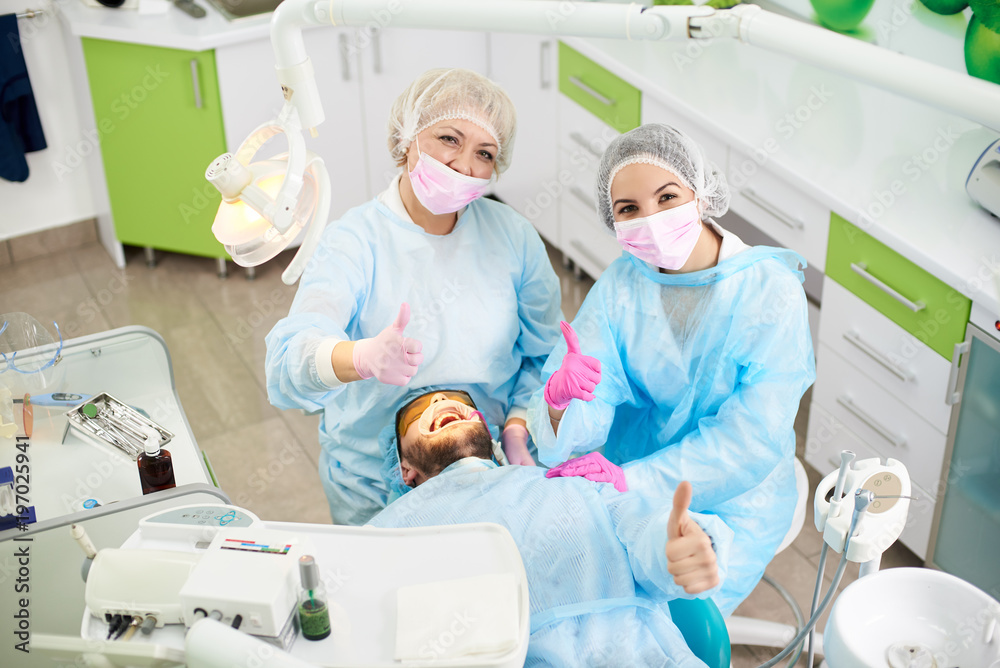 Happy and smiling dentists and their male client showing all thumbs up after successful dental treatment in a dental office. Happy and smiling dentists and their male client showing all thumbs up after successful dental treatment in a dental office.