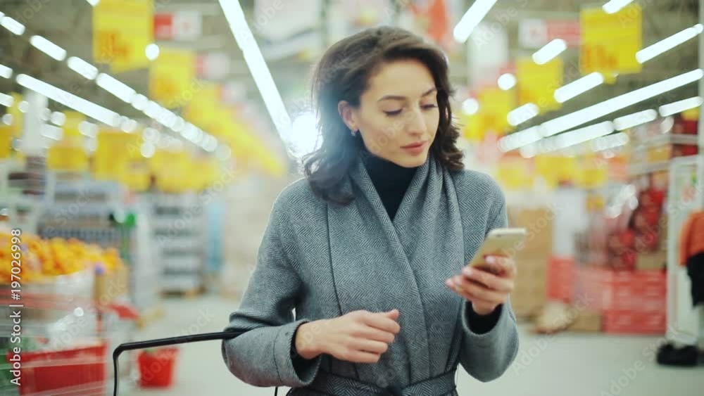 Attractive young woman walk checking to do list on phone in supermarket smiling shopping internet buying technology hypermarket shop mall smartphone shopper grocery market, girl food customer