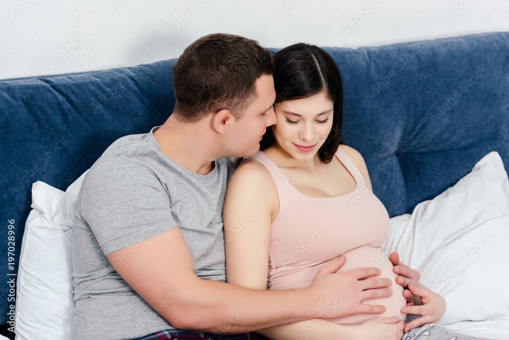 happy young pregnant couple touching belly together on bed happy young pregnant couple touching belly together on bed